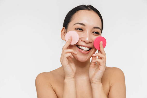Pretty asian woman with healthy skin posing naked isolated over white wall background holding makeup sponge. - Φωτογραφία, εικόνα
