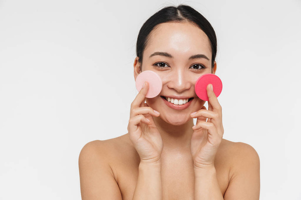 Mooie Aziatische vrouw met een gezonde huid poseren naakt geïsoleerd over witte muur achtergrond Holding make-up spons. - Foto, afbeelding