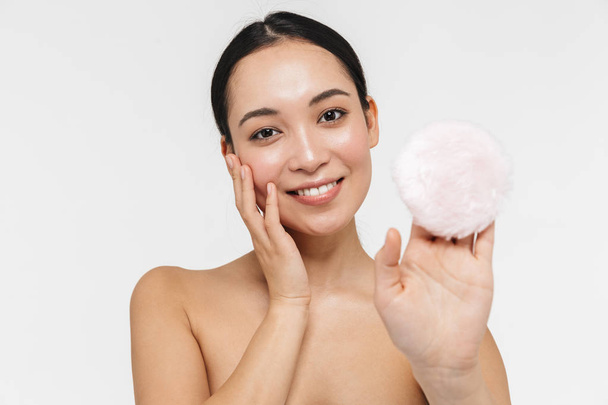 Beautiful young pretty asian woman with healthy skin posing naked isolated over white wall background holding powder puff. - Фото, изображение