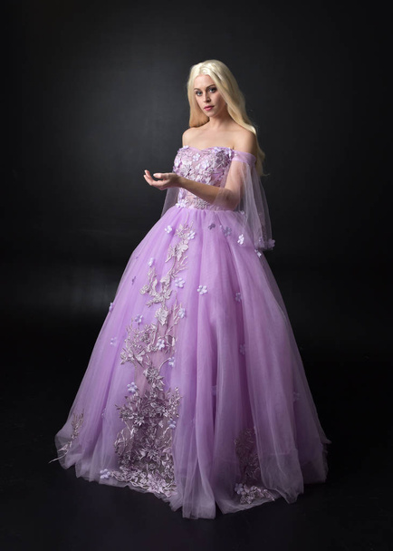 full length portrait of a blonde girl wearing a fantasy fairy inspired costume,  long purple ball gown with fairy wings,   standing pose on a dark studio background. - 写真・画像