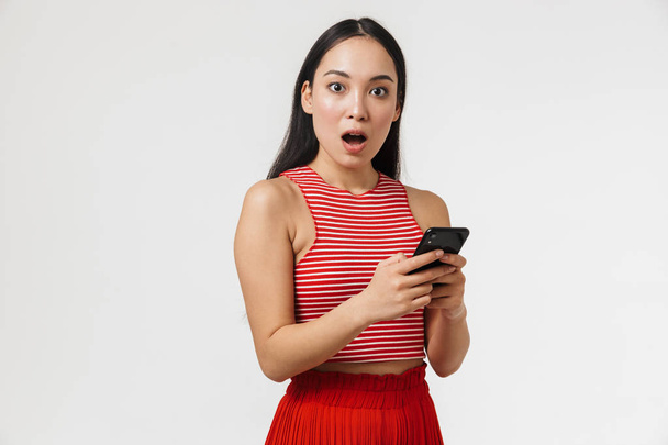 Beautiful young pretty asian shocked woman posing isolated over white wall background using mobile phone. - 写真・画像
