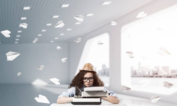 Young woman writer in hat and eyeglasses using typing machine while sitting at the table indoors among flying paper planes and with office view on background. - Photo, Image