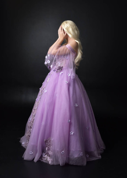 full length portrait of a blonde girl wearing a fantasy fairy inspired costume,  long purple ball gown with fairy wings,   standing pose on a dark studio background. - Photo, Image