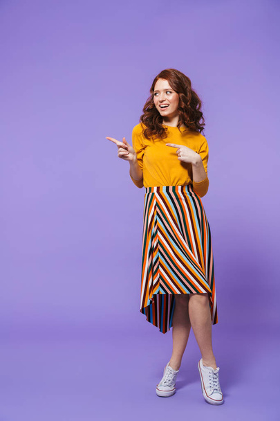 Full length portrait of a pretty young redheaded woman isolated over violet background, pointing finger at copy space - 写真・画像