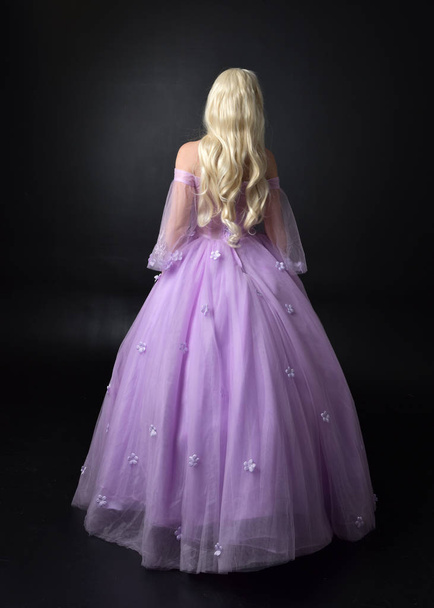 full length portrait of a blonde girl wearing a fantasy fairy inspired costume,  long purple ball gown with fairy wings,   standing pose with back to the camera on a dark studio background. - Photo, Image