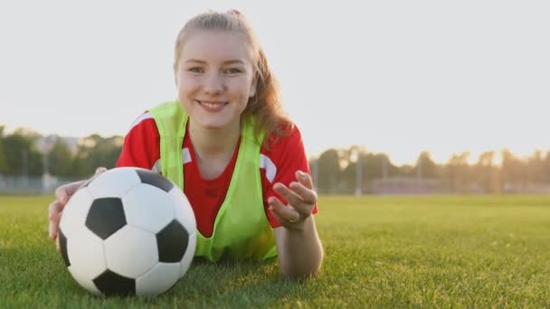 Retrato de una adolescente futbolista sonriente acostada en el campo con pelota de fútbol en cámara lenta
 - Metraje, vídeo