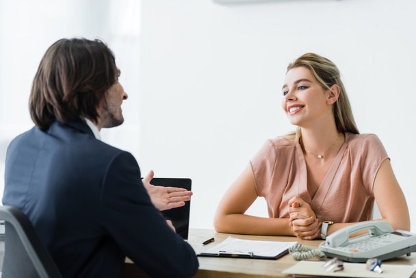χαρούμενη γυναίκα που κάθεται στο γραφείο και μιλάει με τον επιχειρηματία - Φωτογραφία, εικόνα
