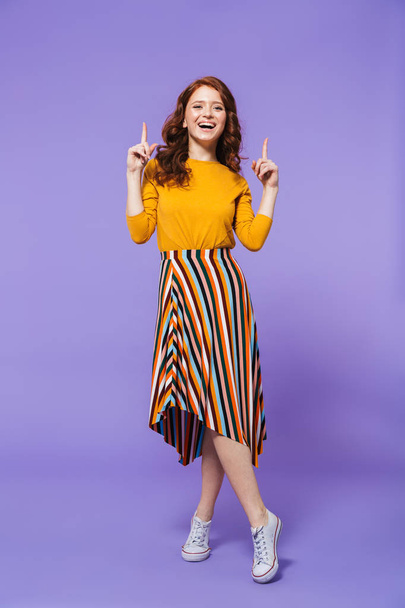 Full length portrait of a pretty young redheaded woman isolated over violet background, pointing finger at copy space - Foto, imagen