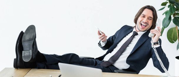 panoramic shot of cheerful businessman talking at telephone, holding smartphone and headset in hands - Φωτογραφία, εικόνα
