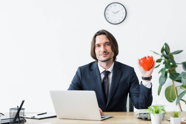 businessman holding red heart in hand, sitting in office and looking at camera, medical insurance concept - Foto, Imagen