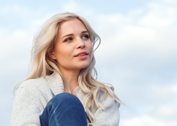Beautiful happy blonde in a gray sweater and jeans smiling against the blue sky. Travel, leisure, tourism concept - Fotó, kép
