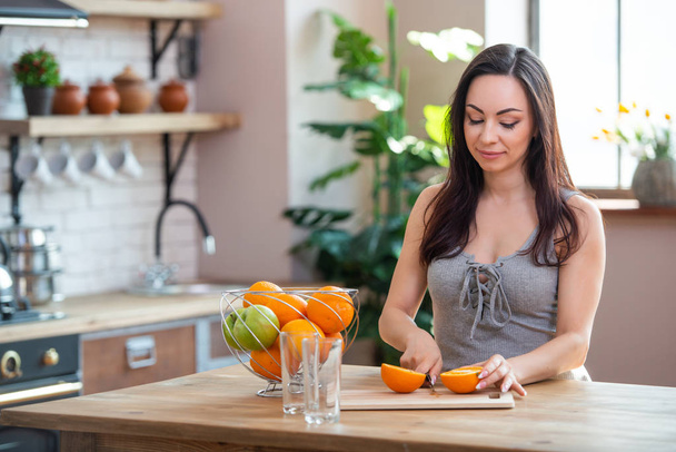 sporty young woman is cutting fresh orange for fruit juice in the kitchen. Horizontal indoors shot - Foto, Imagem
