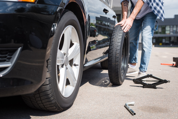 cropped view of man rolling new tire and fixing broken auto, car insurance concept - Фото, изображение