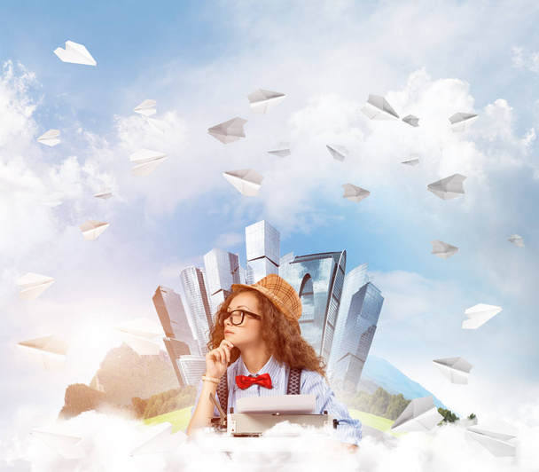 帽子と眼鏡の背景にフローティング市島と曇り空の景色と紙飛行機を飛行中テーブルに座ってタイピング マシンを使用して若い女性作家. - 写真・画像