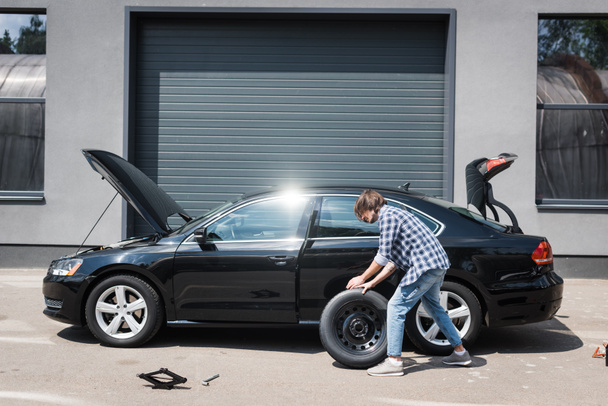 Mann rollt neues Rad und repariert kaputtes Auto in der Nähe von Garage, Kfz-Versicherungskonzept - Foto, Bild