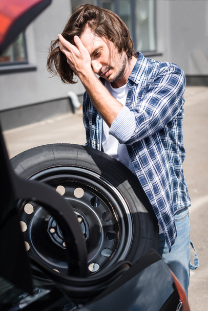 handsome man holding wheel in hand while standing near auto, car insurance concept - Photo, Image