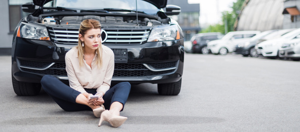 panoramische foto van overstuur zakenvrouw zittend in de buurt van Broken auto met smartphone in handen, autoverzekering concept - Foto, afbeelding
