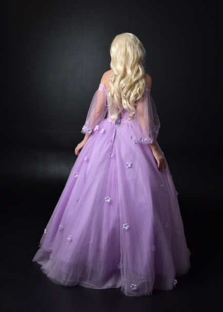 Pełna długość Portret blond dziewczyna ubrana w fantazyjne bajki inspirowane kostium, długa fioletowa suknia balowa z bajki skrzydła, stojący poza z powrotem do aparatu na ciemnym tle Studio. - Zdjęcie, obraz