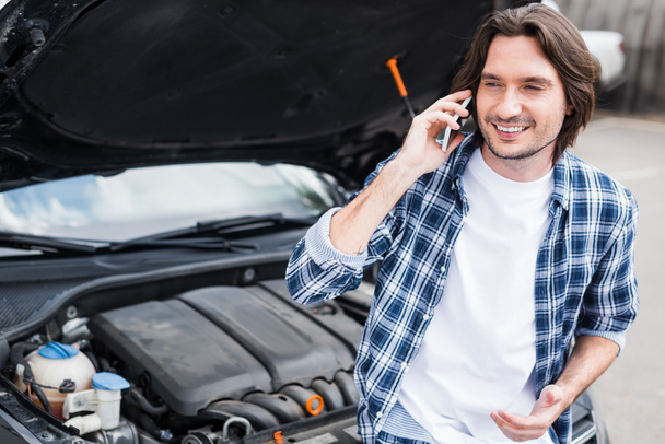 cheerful man in casual cothes talking on smartphone near broken auto with open trunk, car insurance concept - Foto, Bild