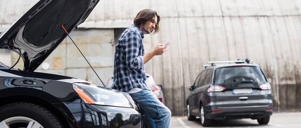 Panoramaaufnahme eines Mannes mit Smartphone, der neben kaputtem Auto mit offenem Kofferraum steht, Kfz-Versicherungskonzept - Foto, Bild