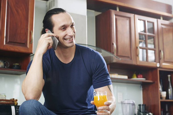 Smiling man sitting and holding orange juice in his hand and laughing during phone call in the kitchen  - Photo, image