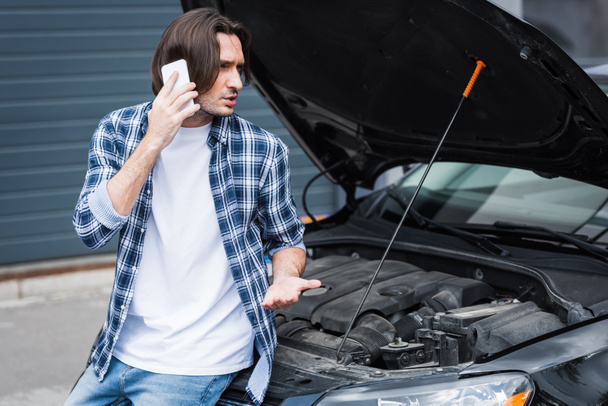 man talking on smartphone while standing near broken auto with open trunk, car insurance concept - Foto, imagen