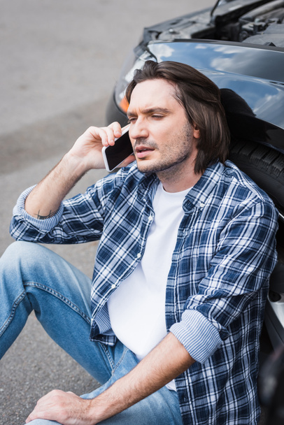 tired man sitting near broken auto and talking on smartphone, car insurance concept - Photo, image