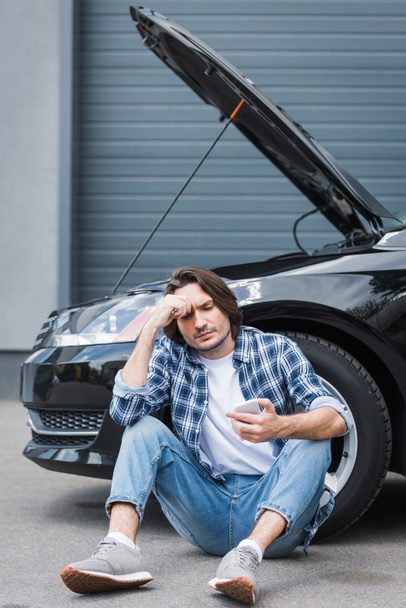 thoughtful man in casual clothes holding smartphone in hand and sitting near broken auto with open trunk, car insurance concept - Fotografie, Obrázek