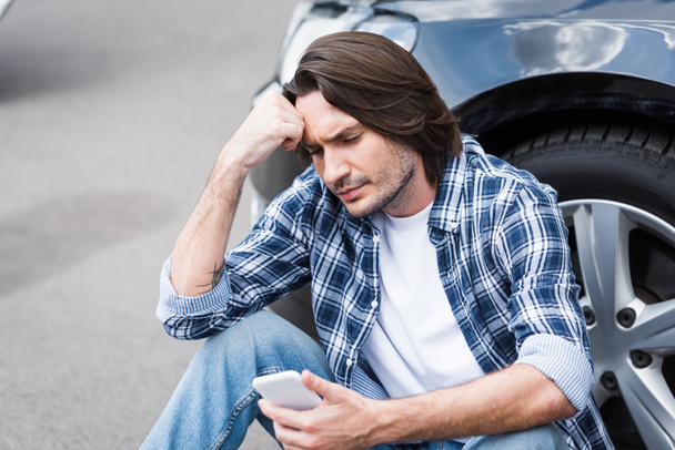 αναστατωμένος άνθρωπος σε casual ρούχα κρατώντας smartphone στο χέρι και καθισμένος κοντά σπασμένο Auto με ανοιχτό κορμό, ασφάλεια αυτοκινήτου concept - Φωτογραφία, εικόνα