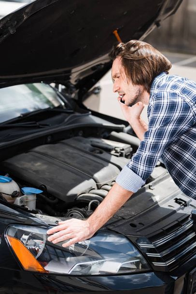 frustrated man talking on smartphone while standing near broken auto with open trunk, car insurance concept - Photo, Image