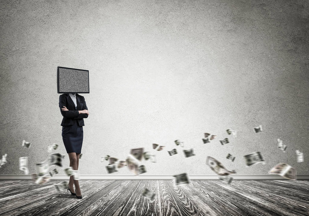 Business woman in suit with TV instead of head keeping arms crossed while standing among flying dollar banknotes inside empty room with gray dark wall on background. - Foto, Imagen