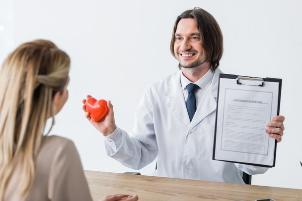 избирательный фокус счастливого врача, держащего красное сердце и страховую форму в руках, глядя на пациента
 - Фото, изображение