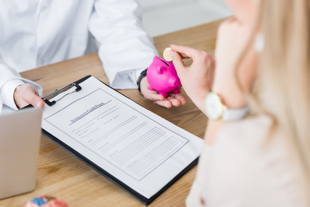 cropped view of doctor holding insurance claim form and piggy bank in hand while woman holding coin - Photo, Image
