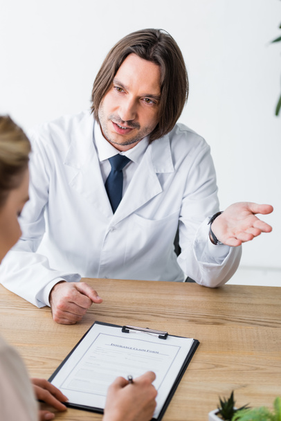 arts praten met patiënt terwijl vrouw ondertekenen document en zittend achter houten tafel - Foto, afbeelding