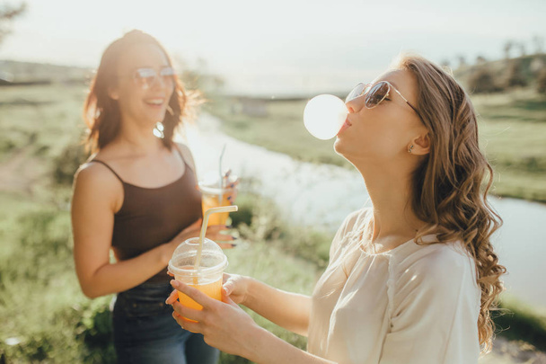 two young girl blowing up a bubble from a chewing gum, drinking orange juice in a plastic cup, at sunset, positive facial expression, outdoor. inflates a chewing gum.  - Foto, imagen