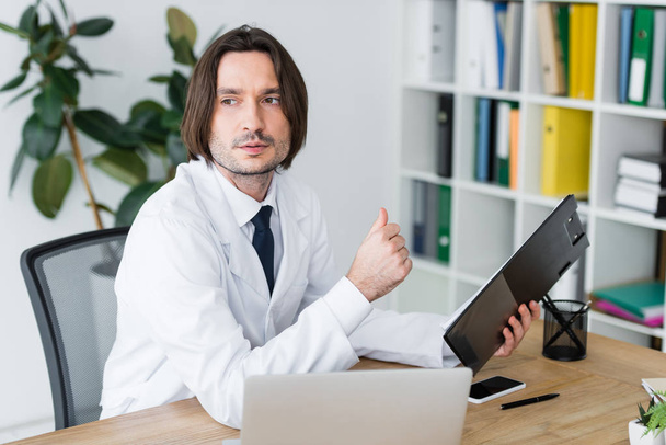 handsome doctor in bright office sitting behind wooden table, looking away and holding clipboard in hand - Fotografie, Obrázek