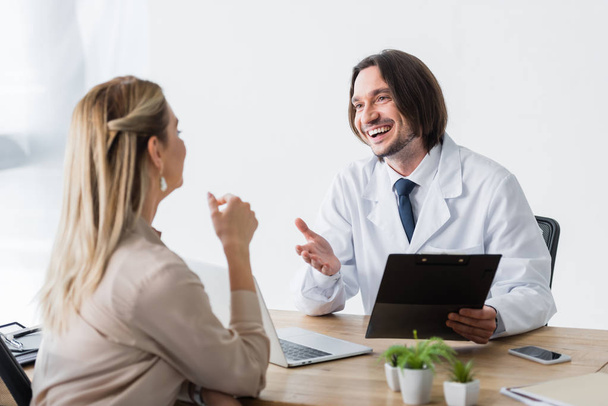 selective focus of happy doctor with clipboard in hand talking with patient - Photo, Image