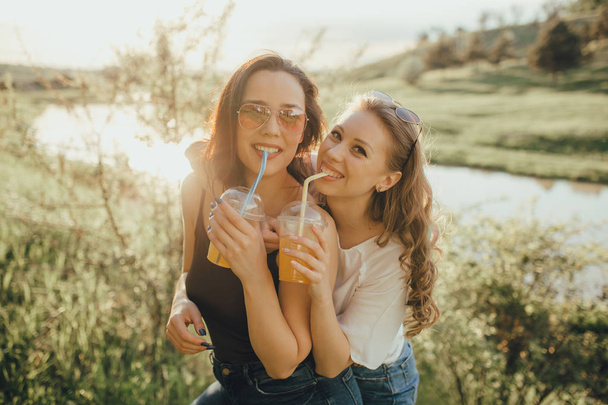 happy girls have fun, drink cocktail from plastic cup with straw in sunglasses, dressed in white and black shirt, at sunset, positive facial expression, outdoor - Фото, изображение