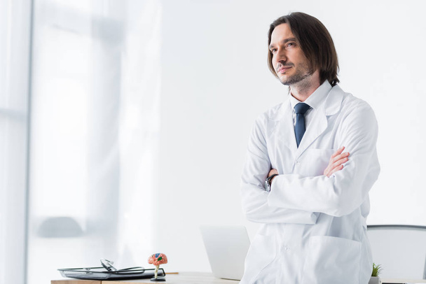 calm doctor in white coat standing in office with crossed hands on chest and looking at camera - 写真・画像
