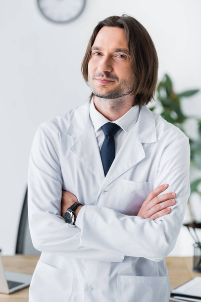 cheerful doctor in white coat standing in office with crossed hands on chest and looking at camera - Fotó, kép