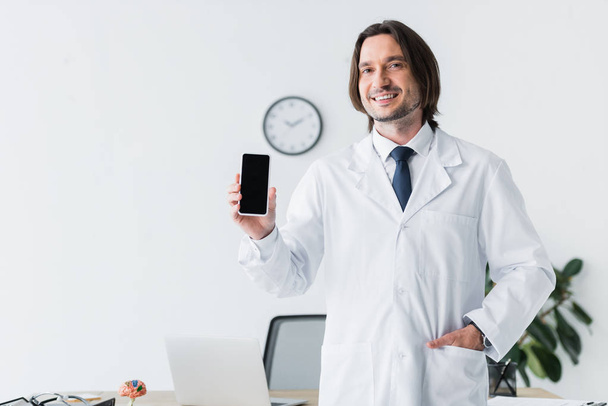 médico alegre en bata blanca mirando a la cámara y sosteniendo el teléfono inteligente en la mano
 - Foto, imagen