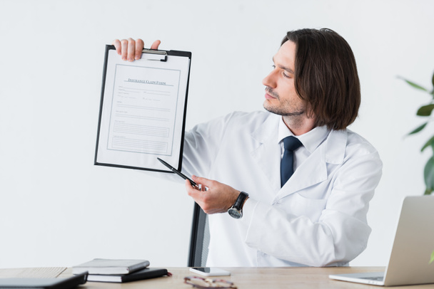 knappe arts in witte vacht wijzend met pen op medisch document  - Foto, afbeelding