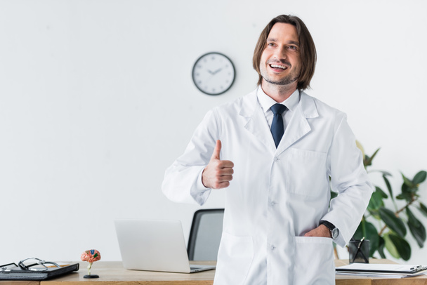 χαρούμενος γιατρός με λευκό παλτό κοιτάζοντας μακριά και δείχνει τον αντίχειρα προς τα πάνω χειρονομία - Φωτογραφία, εικόνα