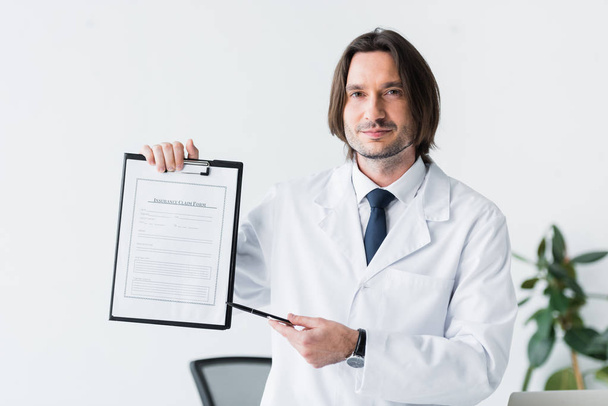 χαμογελαστός γιατρός σε λευκό παλτό κοιτάζοντας την κάμερα και δείχνοντας με στυλό σε ιατρικό έγγραφο  - Φωτογραφία, εικόνα