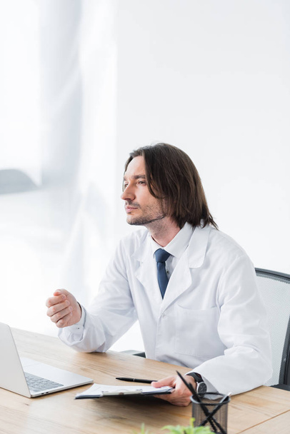 pensive doctor in white coat looking away and sitting behind wooden table in office - Photo, Image