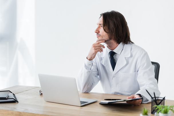 doctor in white coat looking away while sitting behind wooden table with laptop - Fotó, kép