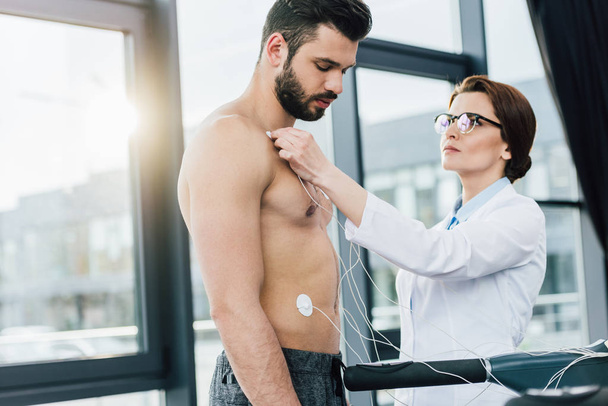 beautiful doctor putting electrodes on shirtless sportsman during endurance test - Foto, imagen