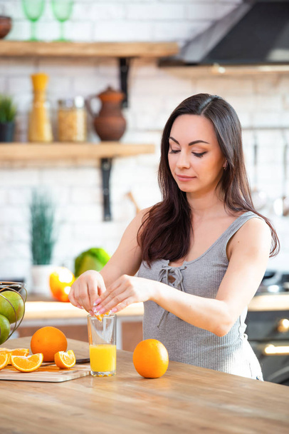 A young woman in a gray T-shirt squeezes a fresh orange on fruit juice. Healthy eating concept. In the interior of the kitchen - Photo, image