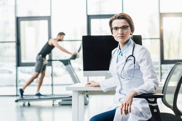 doctor sitting at desk near sportsman running on treadmill during endurance test in gym - Zdjęcie, obraz