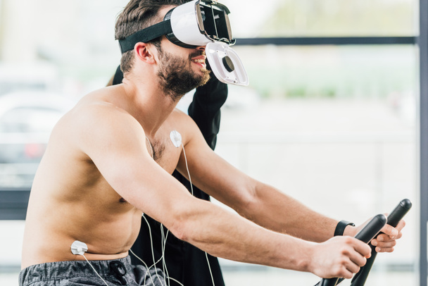 Lächelnder hemdsloser Sportler im Virtual-Reality-Headset mit Elektroden, die während eines Ausdauertests im Fitnessstudio auf Ellipsen trainieren - Foto, Bild